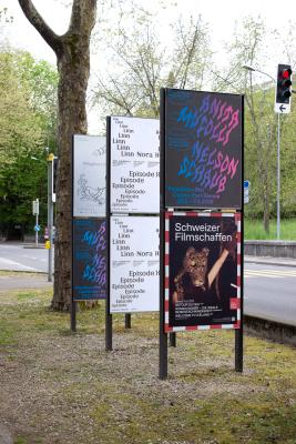 Street photography of an outdoor billboard cluster showing the exhibition poster in context alongside other posters, demonstrating the design's visual presence in a real urban environment.