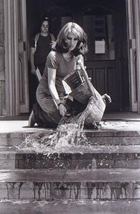 The artist kneels at the top of some steps and pours a bucket of water down them in order to clean.