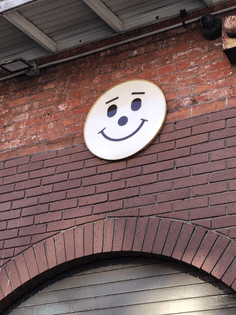 red brick wall with white circle containing two oval eyes one circle nose one curved line smile and two lines as eyebrows  