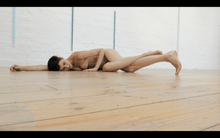 a white person with short brown hair lies naked on wooden floors with white walls. one arm is stretched out above them on the floor and their legs wrapped around each other. 