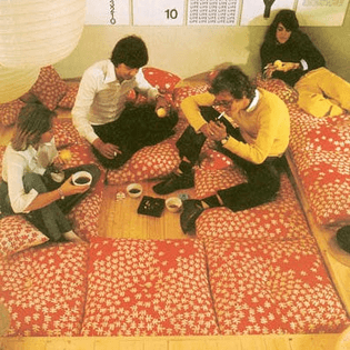 four people sit on square mats arranged on the floor (like tatami), with a blank space in the middle uncovered — to place bowls, and an ashtray. a paper light/lantern is pictured in the upper right corner; it hangs quite low.
