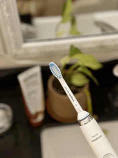 A picture of an electronic toothbrush with toothpaste, a small plant, a mirror, and sink in the background.