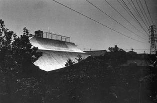 Monochrome photo of a house designed by Shinohara Kazuo, with an undulating roof ducking beneath a large electricity tower and its lines.