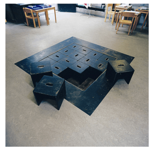 16 stools that fit within a square sunken in the floor of a montessori school classroom. Three of the stools have been taken out to demonstrate how the stools and storage function.
