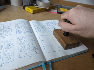 Image of a custom rubber stamp for making thumbnail grids, blue outlined lines with a grid of 2 by 3. 4 of them fit in an A5 sketchbook
