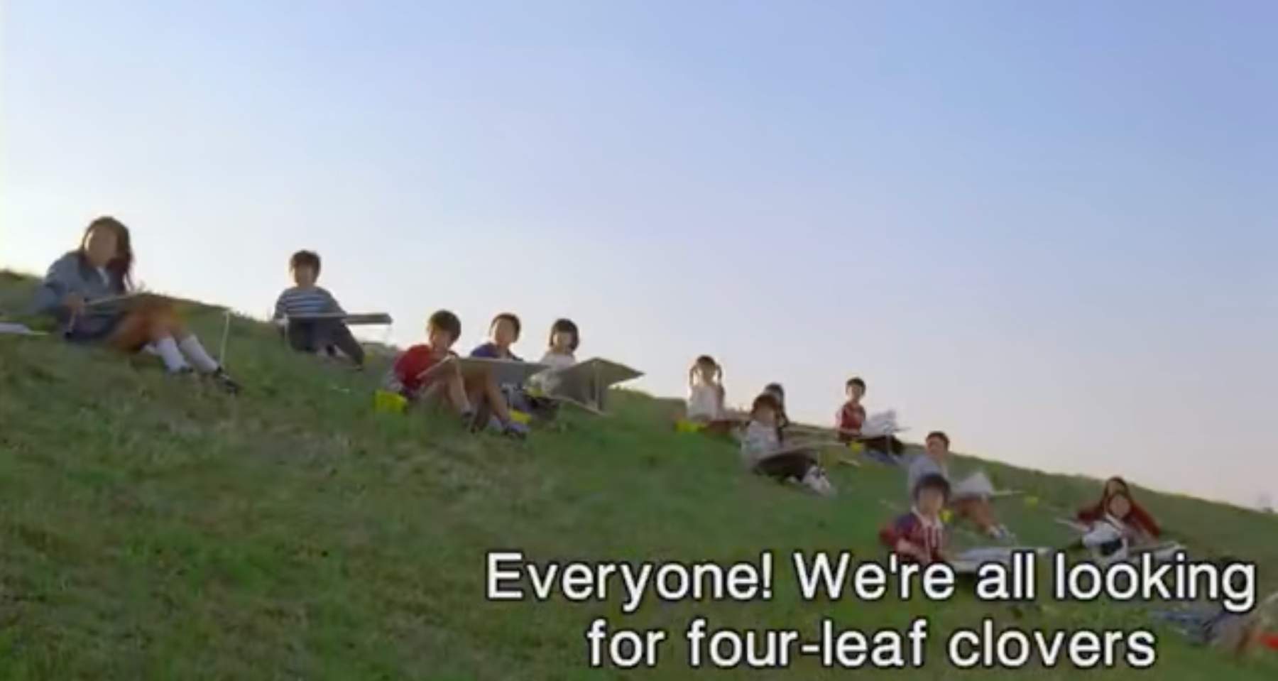 A screencap from Honey and Clove (2006) of children sitting on a grassy mound holding large sketchbooks as part of an art class. The subtitle at the bottom reads "Everyone! We're all looking for four-leaf clovers"