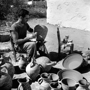 photo of a greek man from skyros painting ceramics somewhen between 1945 and 1955 with a kitty sitting on a little chair next to him (by voula papaioannou)