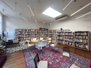 An image of the cybernetics library collection in the meditation room of the Prime Produce Guild Hall. two walls of a square room are lined with bookshleves. a long table with chairs sits on a large red rug, in front of which are two floor chairs. to the left of the long table is a flat file with some containers filled with zines and other matierals. There is a skylight in the middle of the ceiling illuminating the space.